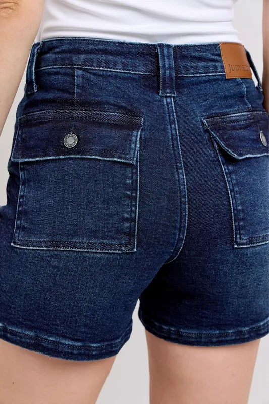 Close-up: dark denim shorts with back flap pockets and tan logo patch